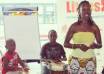Black female teacher in yellow top and African skirt presenting with two child drummers behind her