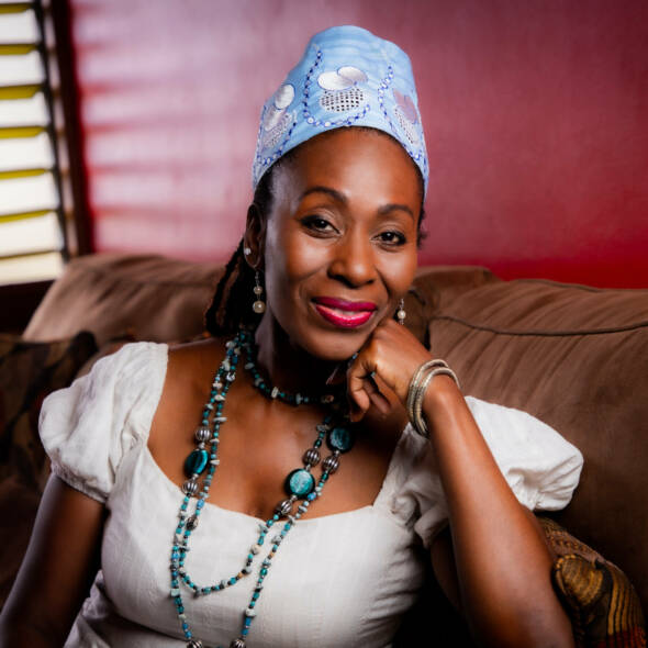 Confident black woman with left arm bent and hand on chin. Lady is white top with mixed colour white and blue beads and blue head wrap
