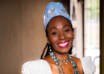 Black woman in white African top and blue hat and skirt smiling with hands on hips